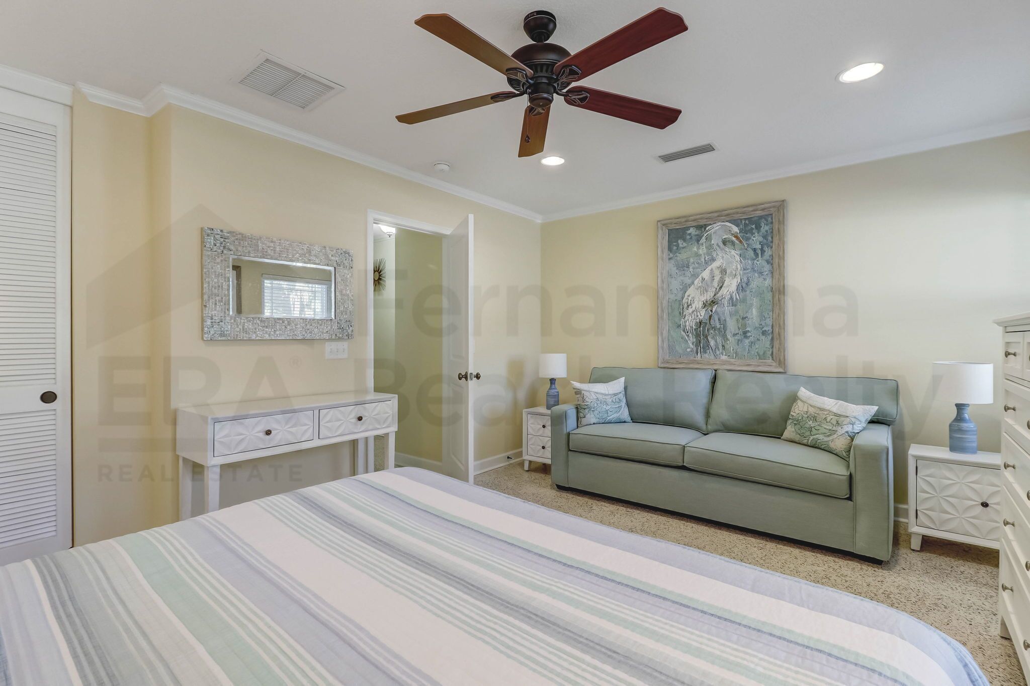 A bedroom with a bed, a green sofa, a ceiling fan, a painting, and a dresser.