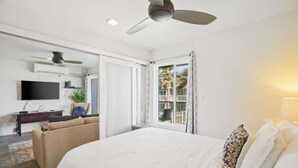Une chambre à coucher avec un lit, un ventilateur de plafond, une télévision, un bureau et une fenêtre avec des rideaux.