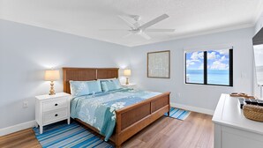 A bedroom with a wooden bed, a ceiling fan, a nightstand with lamps, a window with a view of the ocean, and a blue striped rug. - ✨ Updated Direct Oceanfront Corner 2/2 | Large Wrap-Around Balcony 🌅 | Heated Pool (New Smyrna Beach)