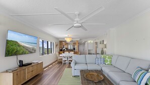 A modern living room with a large flat-screen TV, a ceiling fan, a dining area with a table and chairs, and a kitchen in the background. - ✨ Updated Direct Oceanfront Corner 2/2 | Large Wrap-Around Balcony 🌅 | Heated Pool (New Smyrna Beach)