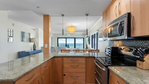 A modern kitchen with wooden cabinets, granite countertops, a stainless steel microwave, and a view to the outside. - ✨ Updated Direct Oceanfront Corner 2/2 | Large Wrap-Around Balcony 🌅 | Heated Pool (New Smyrna Beach)