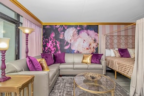 A modern hotel room with a sectional sofa, a round glass coffee table, a bed with a patterned bedspread, and a large floral mural on the wall.