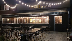Un área de estar al aire libre con mesas y sillas, un letrero que dice "TAVERN ON DIVISION" y secciones que indican "STEAKS", "CHOPS", "SEAFOOD", "PASTA" y "COCKTAILS".