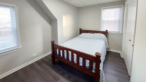 Un dormitorio con una cama de madera, dos ventanas con persianas, techo inclinado y una lámpara.