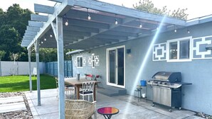 Eine überdachte Terrasse mit Tisch und Stühlen, einem Grill und einer Pergola mit Lichterketten.