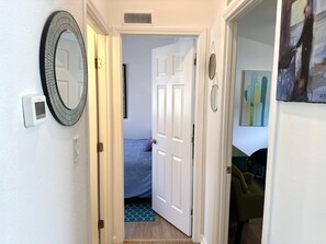 Un couloir avec une porte blanche donnant sur une chambre à coucher où se trouvent un lit et un tapis bleu. Un miroir est accroché au mur et un thermostat est fixé sur le côté gauche.