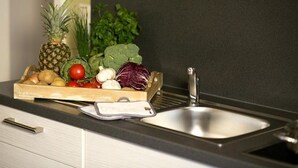 Un plan de travail de cuisine avec une caisse en bois contenant des légumes et un ananas, un évier en acier inoxydable et une balance blanche.