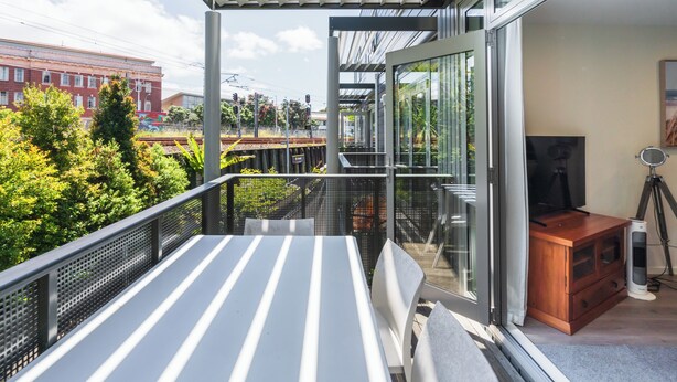 A balcony with a table and chairs, a wooden cabinet with a television, and a glass door leading to a garden. - Spacious Home Near CBD | SPA Pool | Parking | 3BR (Auckland)