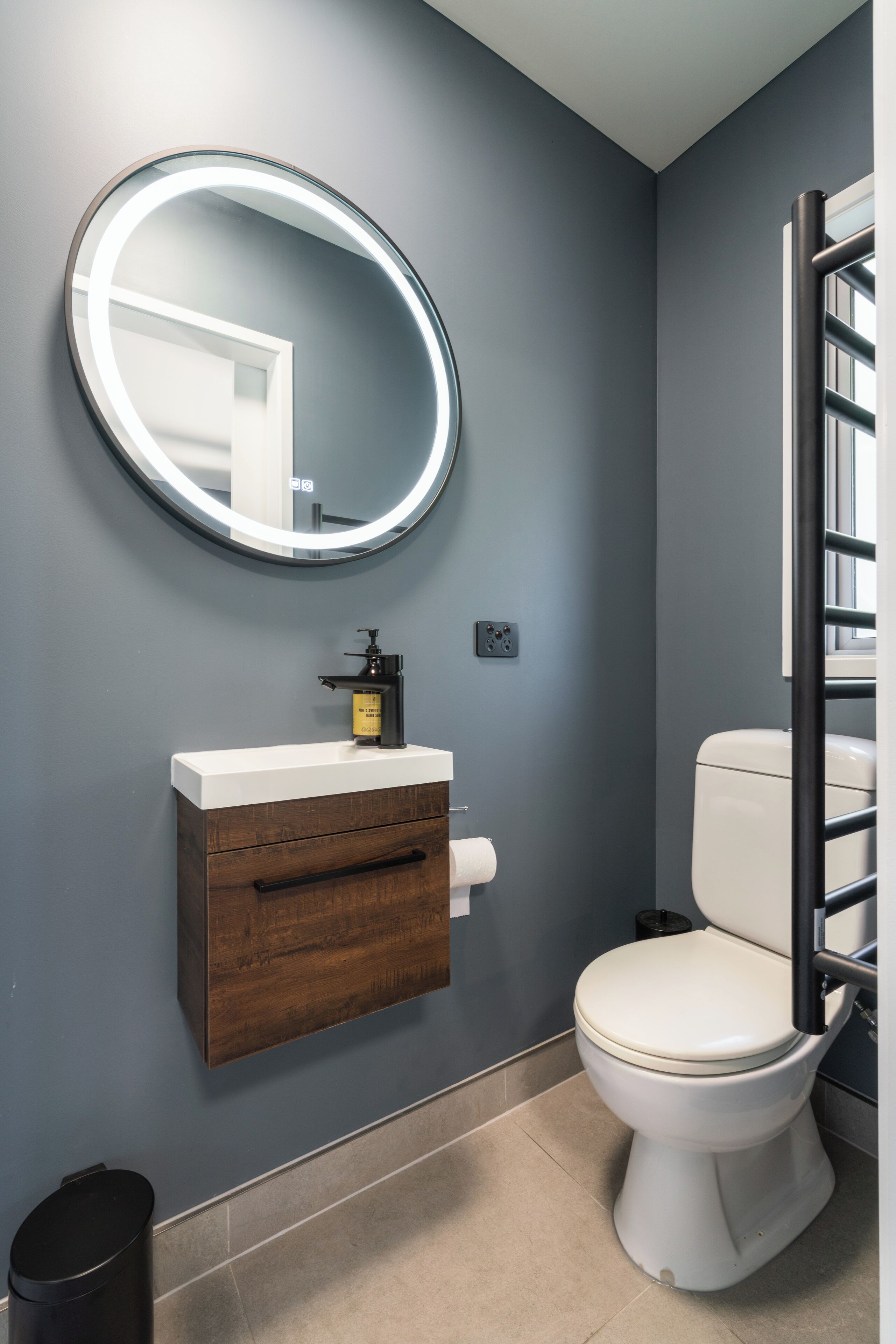 A bathroom with a round mirror, a wooden vanity, a toilet, and a window with a metal railing.