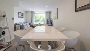 A modern dining area with a white table and chairs, a sofa in the background, and a view of greenery outside. - Spacious Home Near CBD | SPA Pool | Parking | 3BR (Auckland)