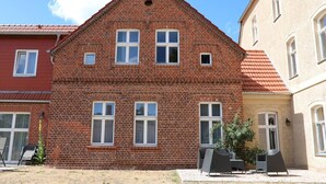 Un bâtiment en briques rouges, doté d’un toit à pignon, de nombreuses fenêtres et d’un petit porche aménagé avec du mobilier d’extérieur.