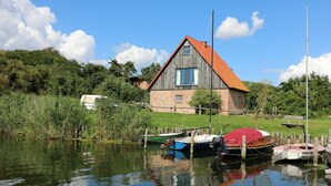 Une maison rustique en bois, avec un toit rouge, située au bord d’une étendue d’eau calme, où se trouvent amarrés quelques bateaux.