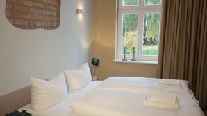 Un lit double avec du linge de lit blanc, un mur en briques apparentes, une lampe fixée au mur, une fenêtre avec des rideaux et une petite table de chevet avec une lampe.