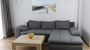 Un salon moderne avec un canapé d’angle gris, une table basse en bois sur laquelle sont disposés des fruits, et un tableau sur le mur représentant un paysage de plage.