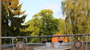 A balcony with a table and two chairs, two glasses of red wine, and a view of trees and a building in the background.