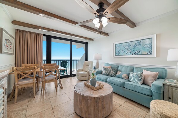 A spacious living room with a large window, a ceiling fan, a teal sofa, and wooden chairs. - Island Winds East 803 | Steps to the HangOut! | Beachfront | My Beach Getaways (Gulf Shores)