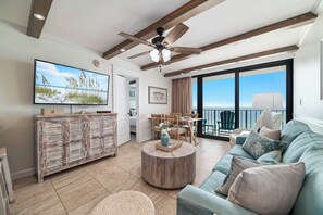 A spacious living room with a large flat-screen TV, a ceiling fan, a wooden coffee table, a sofa, and a balcony with a view. - Island Winds East 803 | Steps to the HangOut! | Beachfront | My Beach Getaways (Gulf Shores)