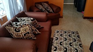 A brown sofa with patterned cushions and a matching ottoman in a room with a tiled floor and a window with curtains.