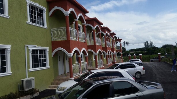 A row of two-story buildings with balconies, parked cars, and a clear sky.