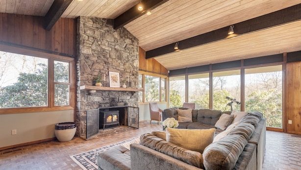A cozy living room with a stone fireplace, a large sofa, and a view of the outdoors. - Asher by AvantStay | Pool + Hot Tub | Stunning Views of the Blue Ridge Mountains (Asheville)