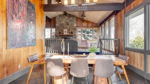 A wooden dining area with a stone fireplace, large windows, and a view of the outdoors. - Asher by AvantStay | Pool + Hot Tub | Stunning Views of the Blue Ridge Mountains (Asheville)