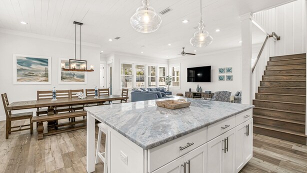 A spacious kitchen with a marble island, wooden dining table, and benches. There is a staircase with wooden steps and a ceiling fan.