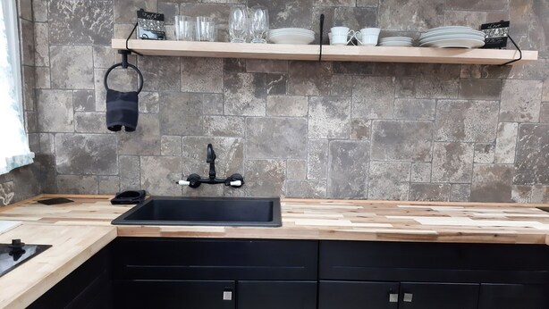 A kitchen with a black sink, wooden countertop, and black cabinets. There is a shelf above the sink holding plates and glasses.