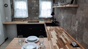 A modern kitchen with a wooden countertop, a built-in sink, and a stone backsplash. There are two windows with floral curtains, a shelf with kitchenware, and a dining table set with plates and cutlery.
