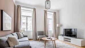 Ein modernes Wohnzimmer mit einem grauen Sofa, einem runden Couchtisch, einem Fernseher auf einem weißen Schrank und zwei Fenstern mit Vorhängen.