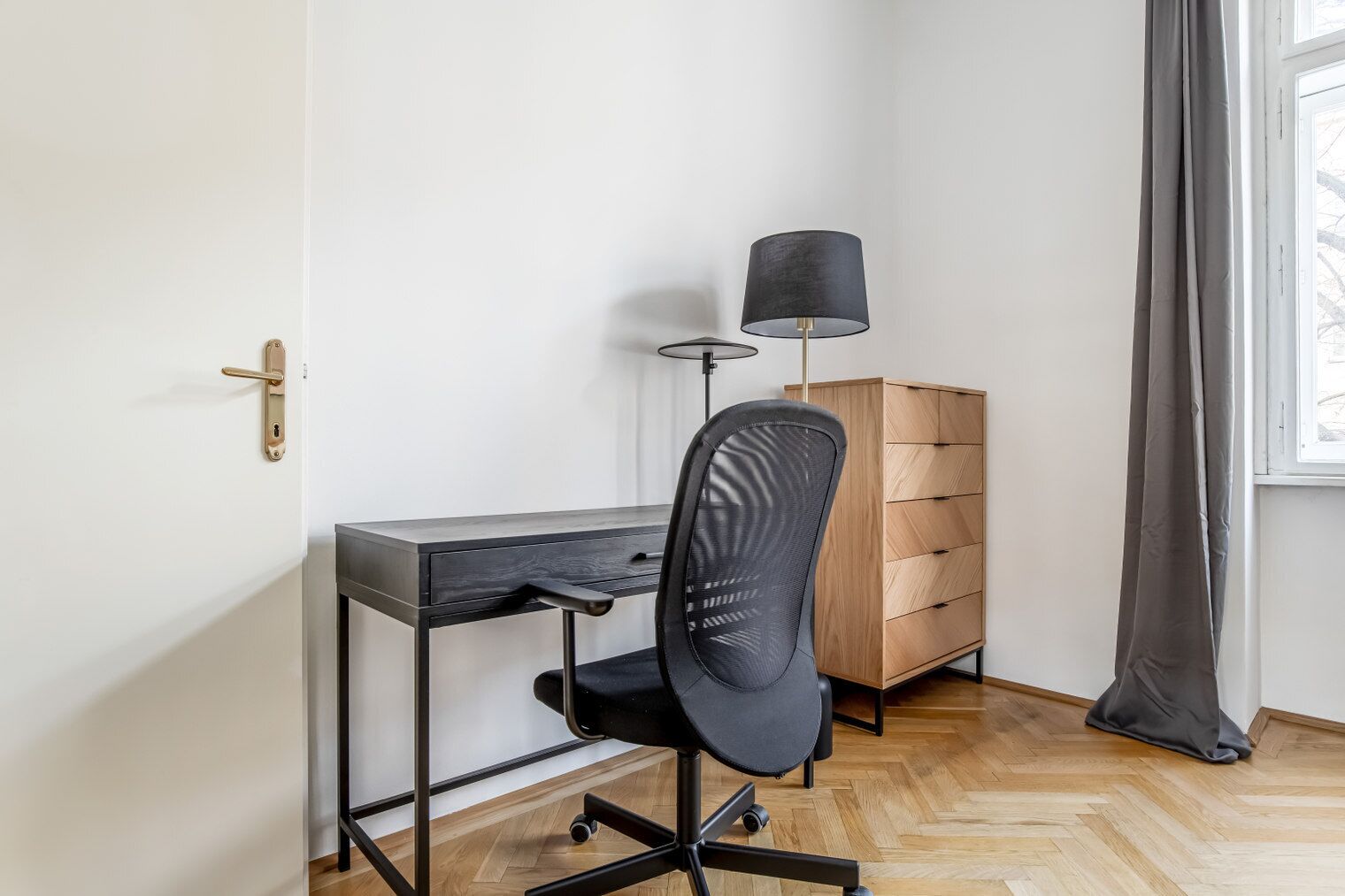Een kleine, moderne kamer met een bureau, stoel, lamp en houten dressoir.