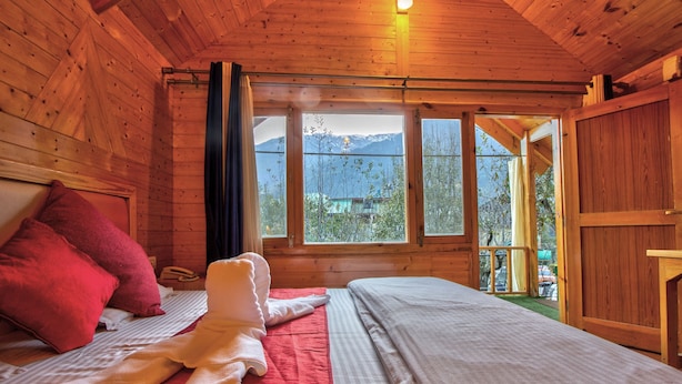 A wooden cabin room with a bed, a window with a view of mountains, and a wooden door. - Attic Cottage by Dumnu Homes (Manali)