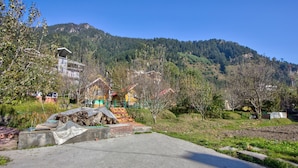 A residential area with houses, a paved road, and a forested hill in the background. - Attic Cottage by Dumnu Homes (Manali)