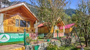 A wooden cabin with a stone wall and a red roof, surrounded by greenery and a mountain backdrop. - Attic Cottage by Dumnu Homes (Manali)