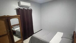 Ein kleiner, gut beleuchteter Raum mit einem Bett, einem Sessel, einem kleinen Tisch und einer Klimaanlage an der Wand.