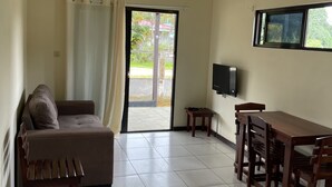 Ein Zimmer mit einer Couch, einem Esstisch mit Stühlen, einem an der Wand befestigten Fernseher und einer Schiebeglastür, die zu einem Außenbereich führt.