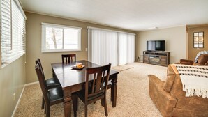 A spacious living room with a dining area, a brown leather couch, a flat-screen TV, and a large window with blinds. - Manistique Lake Retreat w/ Dock! (Germfask)
