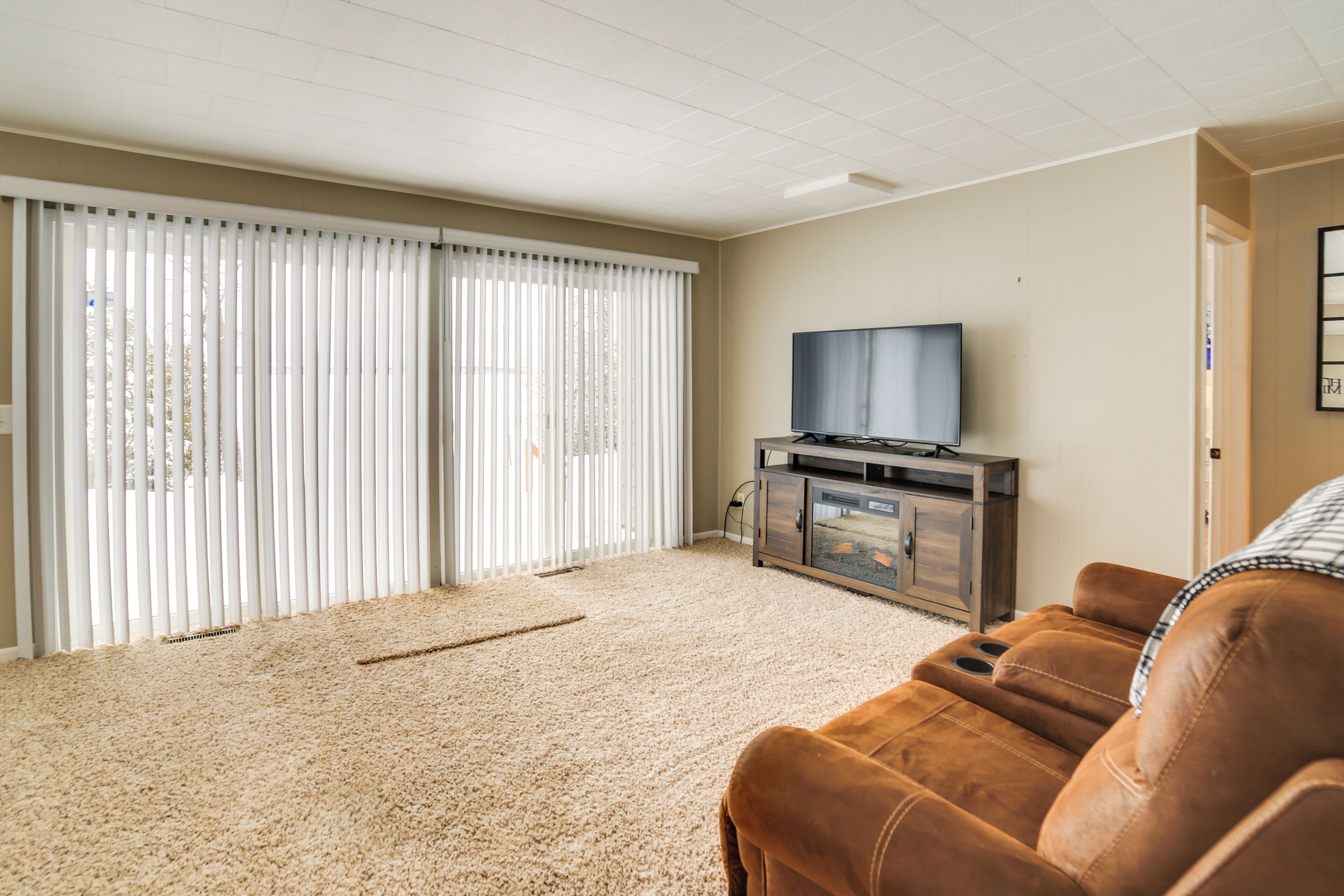 Un salon avec un tapis beige, un canapé en cuir marron, un téléviseur posé sur un meuble en bois et une porte-fenêtre coulissante avec des stores blancs.