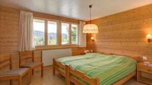 A wooden cabin bedroom with a bed, a chair, a nightstand, and a window with curtains. - Apartment Adele by Interhome (Fiesch)