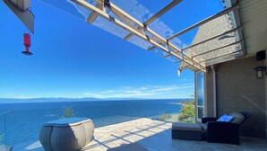 A sunlit outdoor seating area with a view of the sea, featuring wicker furniture and a glass balustrade. - Oceanfront Gated Villa with pool & Hot tub in Nanaimo (Nanaimo)