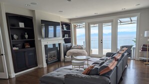Un salon moderne avec un canapé d’angle gris, une cheminée et une grande fenêtre donnant sur la mer.