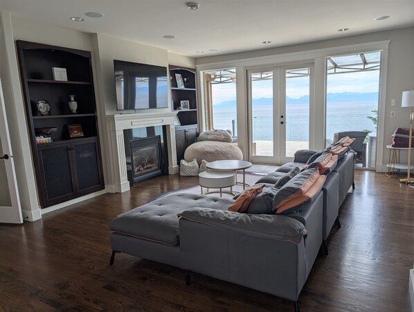 A modern living room with a grey sectional sofa, a fireplace, and a large window offering a view of the sea. - Oceanfront Gated Villa with pool & Hot tub in Nanaimo (Nanaimo)