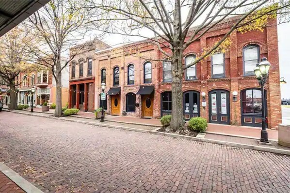 A historic brick building with multiple storefronts, a cobblestone street, and trees with autumn leaves. - Luxury Downtown Paducah 2 Bedroom 2 Bath Double Condo (Paducah)