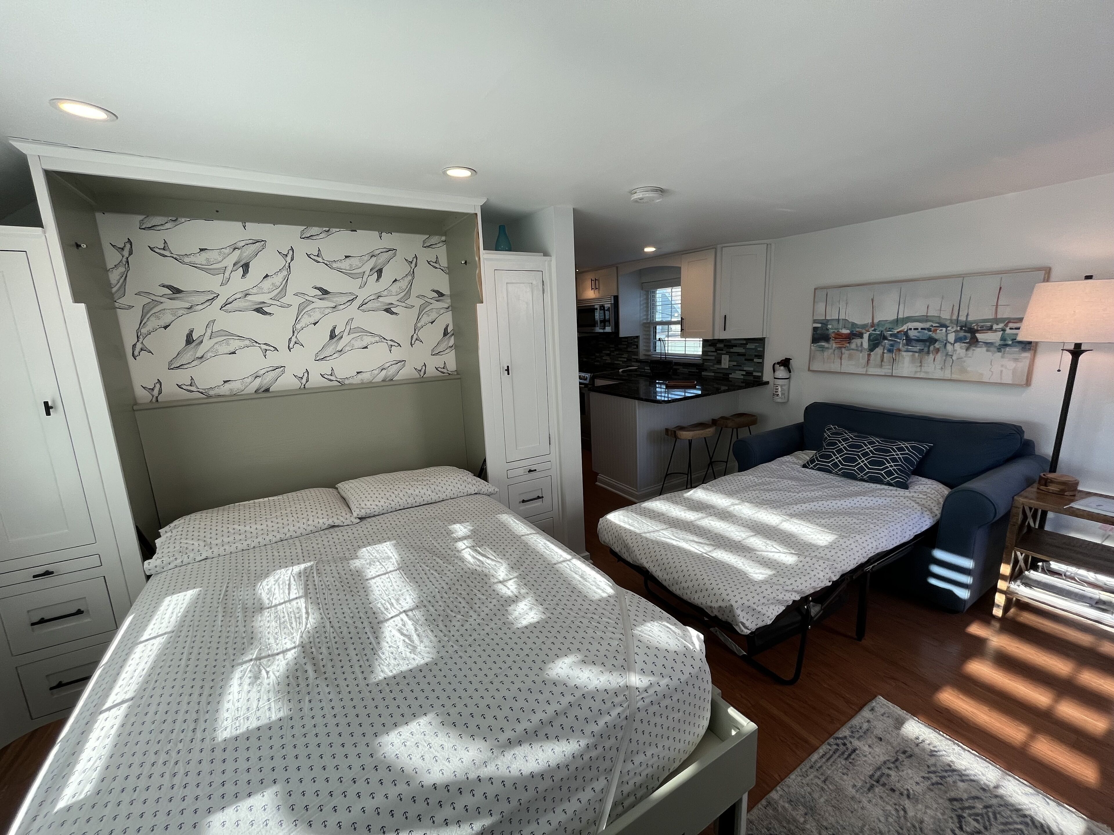 A bedroom with a bed, a sofa, a desk, and a kitchenette in the background.