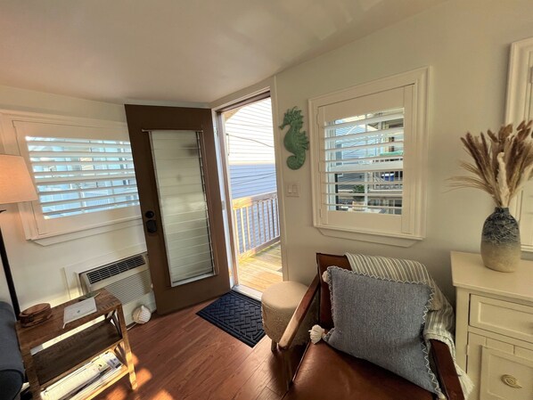 A cozy room with a wooden floor, a brown leather sofa, a small wooden side table, a white dresser, and a large window with blinds.