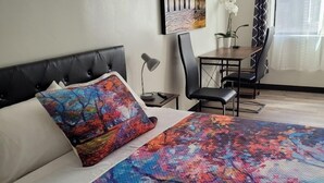 Habitación de hotel con cama, escritorio, silla, planta en maceta y un cuadro en la pared.
