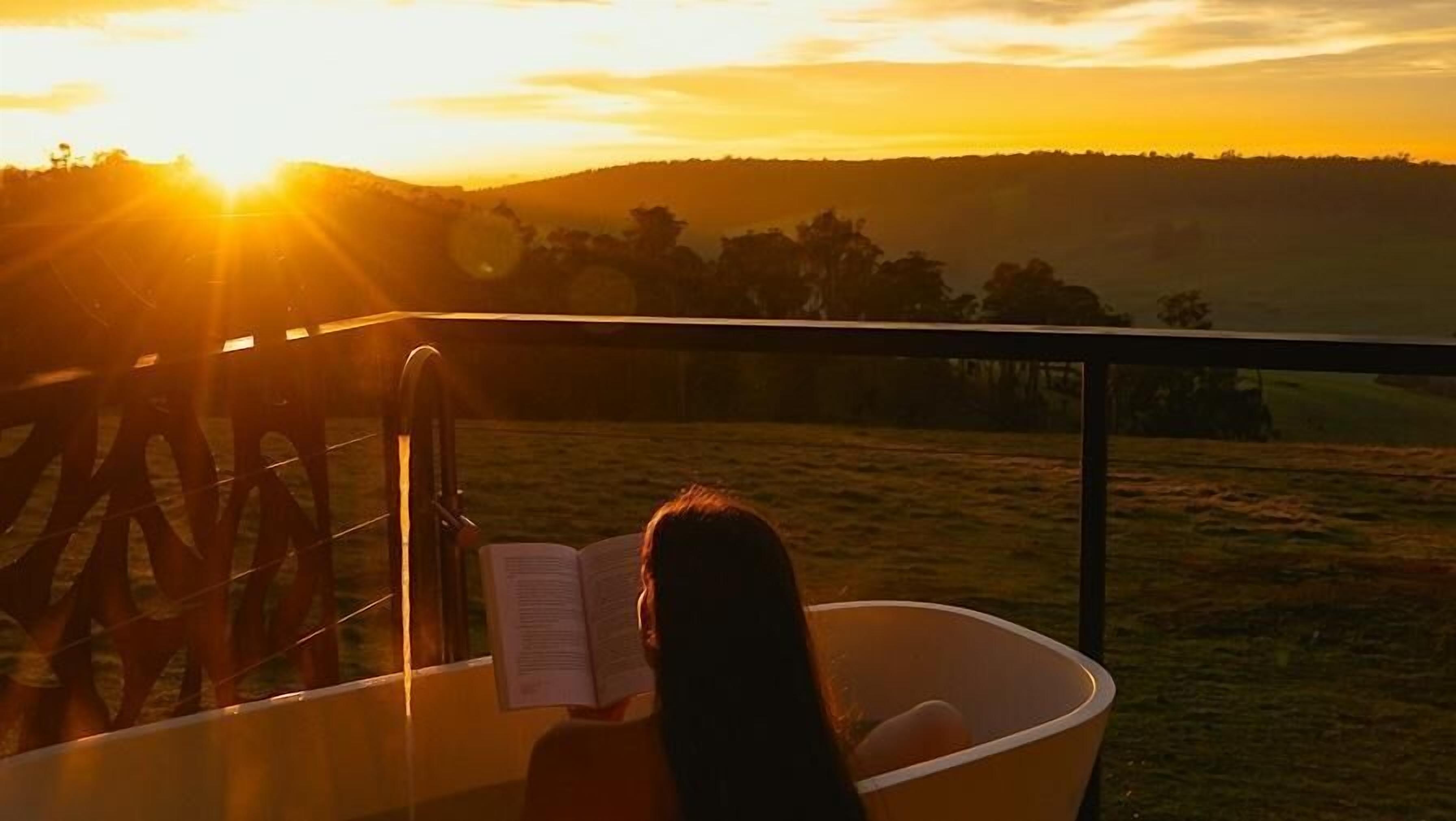 Eine Person liest ein Buch an einem Tisch mit Blick auf einen Sonnenuntergang über einer hügeligen Landschaft.