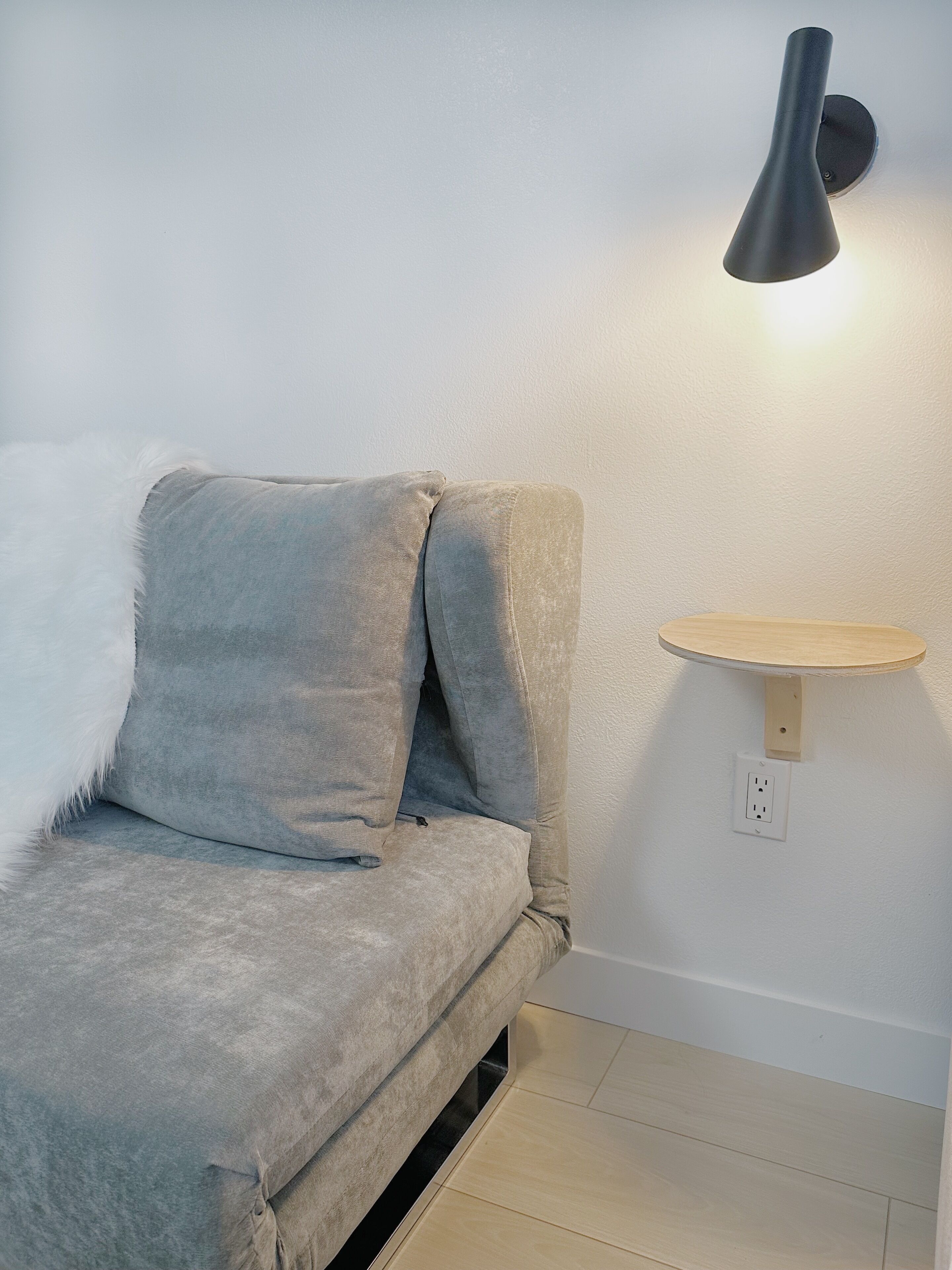 A room with a grey sofa, a wall-mounted light, and a small wooden shelf.