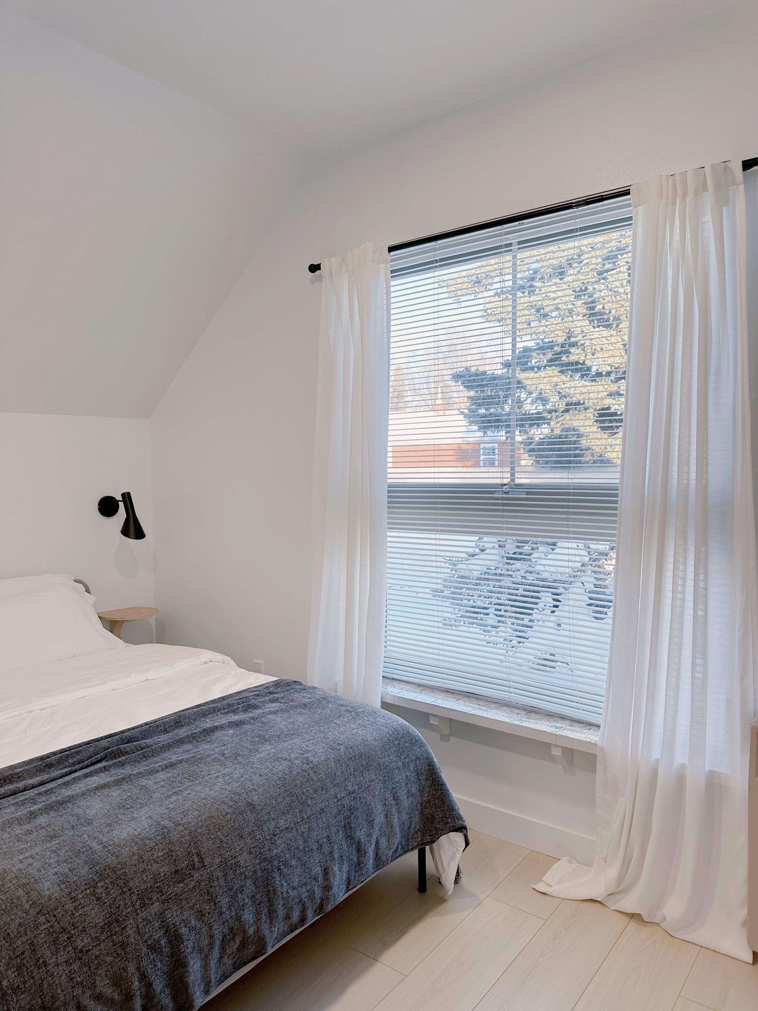 A bedroom with a bed, a bedside table, a lamp, and a window with blinds and curtains.