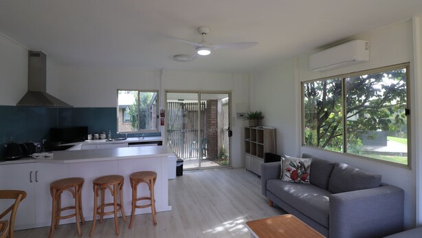 A modern kitchen with a central island, bar stools, a grey sofa, and a wooden table. - Cedric Cottage, 1B Cedric Court (Mallacoota)