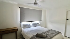 A bedroom with a bed, a ceiling fan, a window with blinds, a bedside table, and a door. - Cedric Cottage, 1B Cedric Court (Mallacoota)